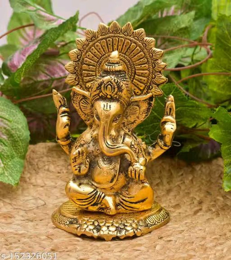 May include: A metallic gold-toned statue of the deity Ganesha, depicted in a seated position with an elephant head, large ears, and a trunk curved to the left. The figure features an ornate circular halo behind the head and detailed engravings.