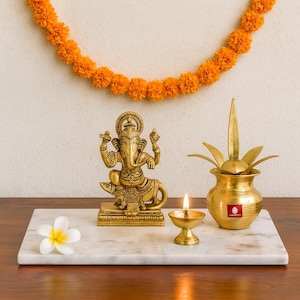 Op de afbeelding: Een gouden beeld van de hindoe-god Ganesha staat op een marmeren dienblad, naast een brandende olielamp en een decoratieve messing pot. Een witte bloem en een slinger van oranje bloemen completeren de opstelling.