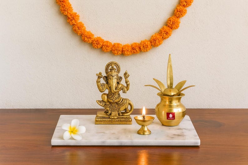 Op de afbeelding: Een gouden beeld van de hindoe-god Ganesha staat op een marmeren dienblad, naast een brandende olielamp en een decoratieve messing pot. Een witte bloem en een slinger van oranje bloemen completeren de opstelling.