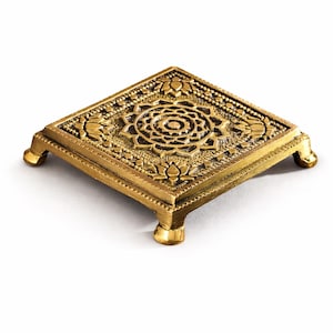 Pode incluir: Um suporte decorativo quadrado dourado com intrincados padrões florais e geométricos. O suporte tem quatro pernas curtas e uma superfície elevada e detalhada. O design apresenta um motivo circular central. O suporte é provavelmente de metal.
