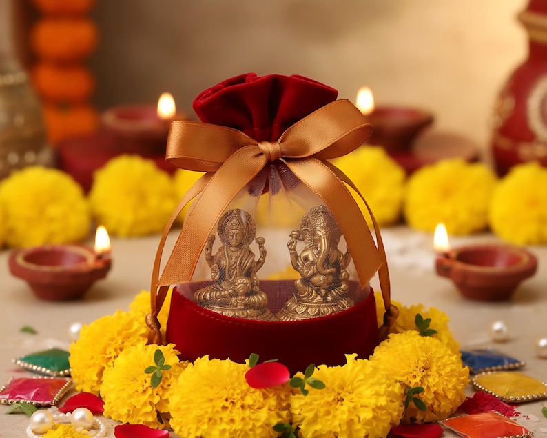Pode incluir: Um conjunto de presentes decorativo com duas pequenas est&aacute;tuas douradas de divindades hindus, fechadas numa bolsa de veludo vermelho amarrada com uma fita dourada. O conjunto &eacute; rodeado por flores de cal&ecirc;ndula amarelas, velas e pequenas l&acirc;mpadas de barro.