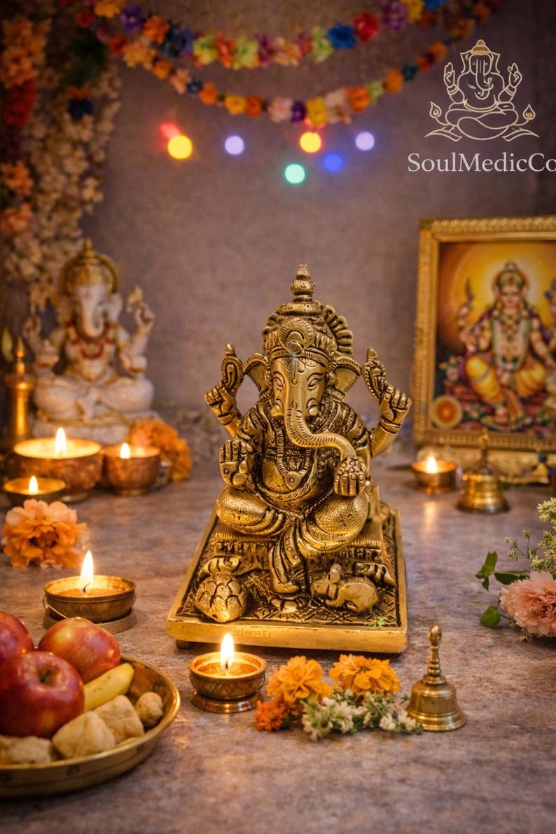 May include: A golden Ganesh statue is the focal point of a spiritual display, surrounded by lit candles, flowers, and a framed image. A small brass bell and a bowl of fruit add to the ambiance. Colorful lights and a garland are in the background.
