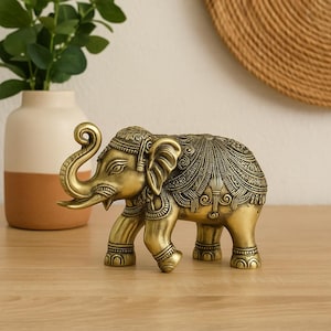 May include: A detailed brass elephant figurine with intricate carvings. The elephant has a raised trunk and is adorned with ornate patterns on its body and head. The figurine is a warm gold color and stands on a wooden surface, suggesting a decorative item.