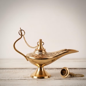 May include: A golden genie lamp with intricate carvings and a curved spout. The lamp features a handle and a removable lid with a chain. The lamp is made of metal and sits on a white wooden surface.