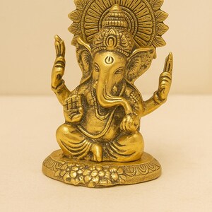 May include: A gold-toned metallic statue of the deity Ganesha in a seated position. The figure features an elephant head, multiple arms, and a large circular halo behind the head. The base is decorated with floral carvings and a smooth finish.