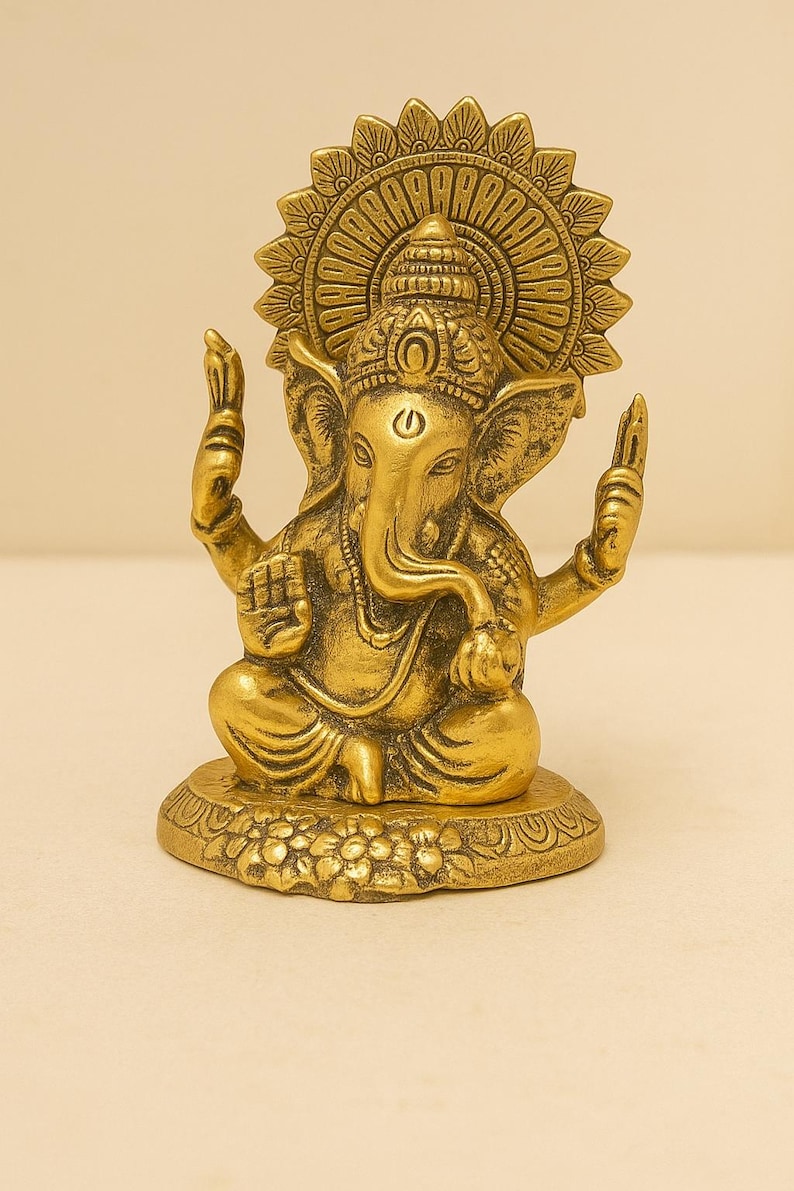 May include: A gold-toned metallic statue of the deity Ganesha in a seated position. The figure features an elephant head, multiple arms, and a large circular halo behind the head. The base is decorated with floral carvings and a smooth finish.