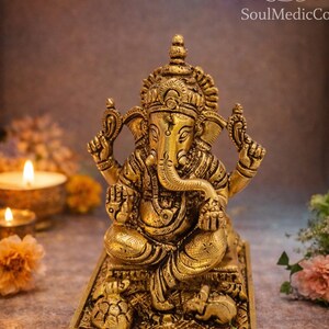 May include: A detailed golden brass statue of the Hindu deity Ganesha, seated on a rectangular base. The statue features intricate carvings and is surrounded by candles and flowers, with the SoulMedicCo logo visible.