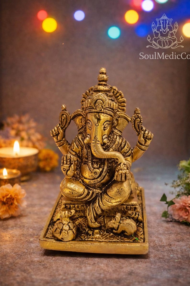 May include: A detailed golden brass statue of the Hindu deity Ganesha, seated on a rectangular base. The statue features intricate carvings and is surrounded by candles and flowers, with the SoulMedicCo logo visible.