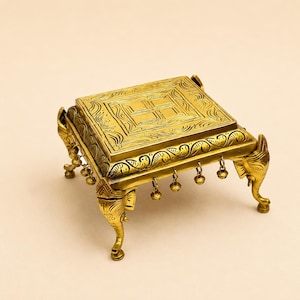 May include: A square brass pedestal stand featuring intricate engraved patterns on the top surface and sides. The stand is supported by four legs shaped like elephant heads and trunks, with small metal bells hanging from the edges of the frame.