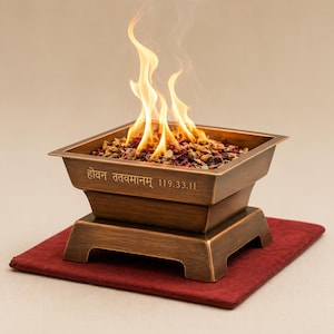 May include: A bronze-colored, square fire pit with flames rising from within. The fire pit sits on a red mat. The text "हवन ततवमानम् 119.33.11" is inscribed on the side. The fire pit is filled with small rocks and dried flowers.
