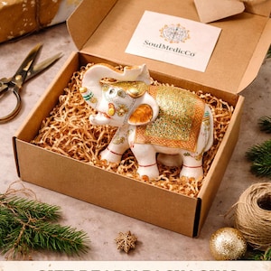 Puede incluir: Una figura de elefante de cer&aacute;mica blanca con detalles dorados y verdes, presentada en una caja de regalo con papel de relleno. La caja est&aacute; abierta, mostrando el elefante, rodeada de decoraciones.