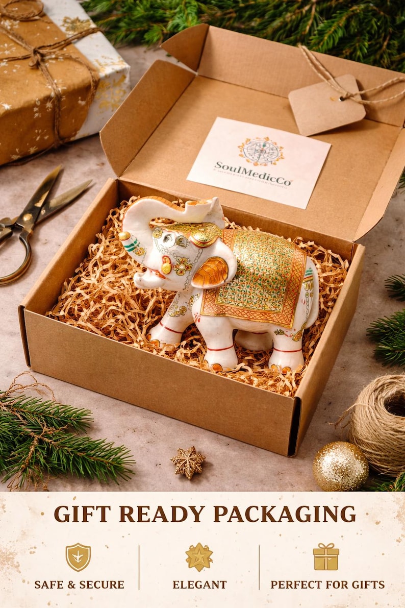 Puede incluir: Una figura de elefante de cer&aacute;mica blanca con detalles dorados y verdes, presentada en una caja de regalo con papel de relleno. La caja est&aacute; abierta, mostrando el elefante, rodeada de decoraciones.