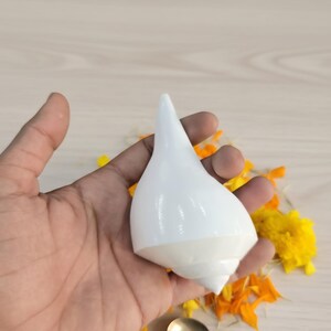 May include: A white conch shell with a pointed top, held in a hand. The shell is surrounded by yellow and orange flower petals. A small, ornate spoon rests below the shell.