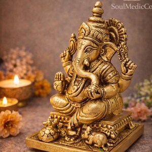 May include: A golden statue of the Hindu deity Ganesha, seated on a square base. The statue features intricate details, including a large elephant head, multiple arms, and ornate clothing. The background includes candles and flowers.