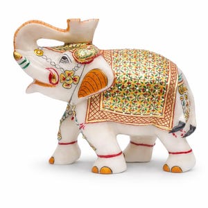 Puede incluir: Una figura decorativa de elefante blanco con intrincados detalles pintados. El elefante presenta detalles en naranja, rojo, verde y dorado en sus orejas, manta y elementos decorativos. La trompa est&aacute; levantada y la figura tiene un dise&ntilde;o ornamentado.