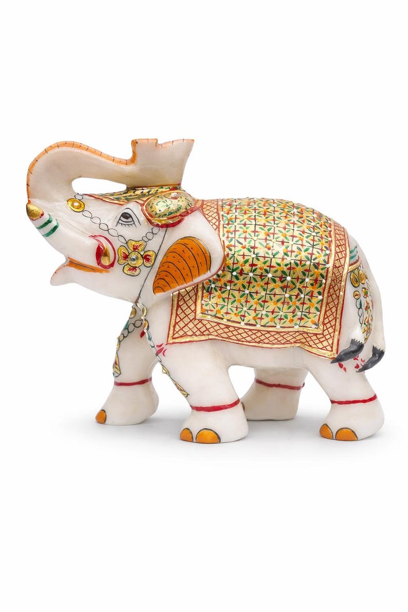 Puede incluir: Una figura decorativa de elefante blanco con intrincados detalles pintados. El elefante presenta detalles en naranja, rojo, verde y dorado en sus orejas, manta y elementos decorativos. La trompa est&aacute; levantada y la figura tiene un dise&ntilde;o ornamentado.