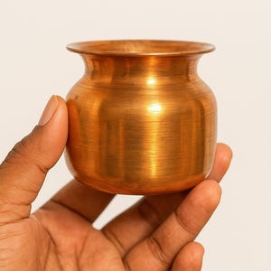 Puede incluir: Una pequeña olla de color cobre con un cuerpo redondeado y un borde ensanchado. La olla se sostiene en una mano sobre un fondo liso. El metal tiene una superficie pulida y reflectante.