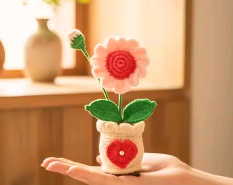 May include: A handmade crochet flower in a small pot. The flower has pink petals and a red center. The pot is beige with a red heart design. Green leaves and a bud are also visible.