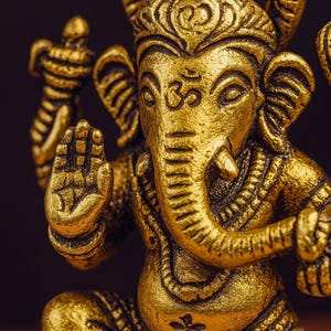 Op de afbeelding: Een gedetailleerd, gouden beeld van de hindoe-godheid Ganesha, met een olifantenkop en meerdere armen. Het beeld is ingewikkeld gesneden, met een serene uitdrukking en een opgeheven hand in een zegenend gebaar. De achtergrond is donker.