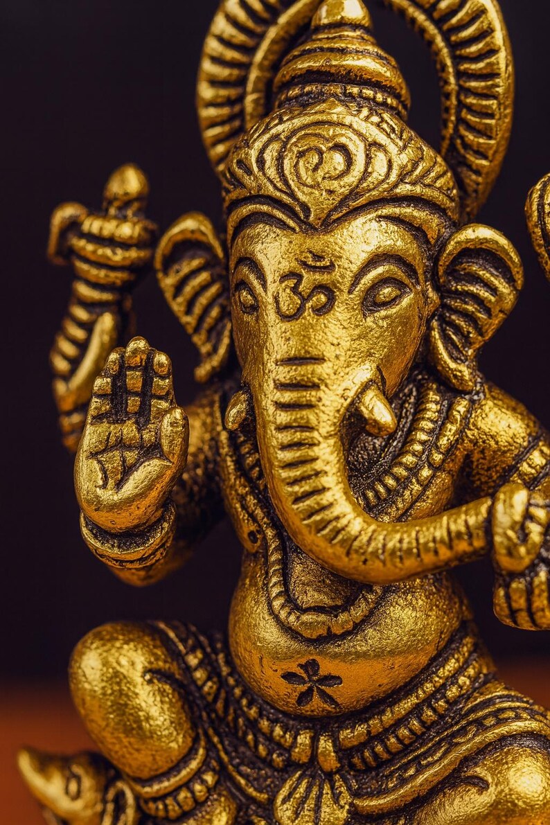 Op de afbeelding: Een gedetailleerd, gouden beeld van de hindoe-godheid Ganesha, met een olifantenkop en meerdere armen. Het beeld is ingewikkeld gesneden, met een serene uitdrukking en een opgeheven hand in een zegenend gebaar. De achtergrond is donker.