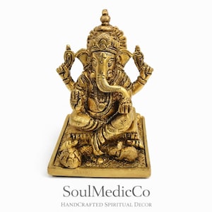 May include: A gold-colored statue of the Hindu deity Ganesha, seated on a rectangular base. The statue features intricate details, including multiple arms, a large elephant head, and a crown. The base has a small inscription and is decorated with small figures.