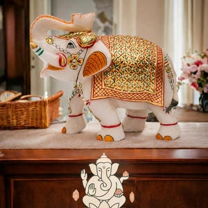 Puede incluir: Una figura de elefante blanco con detalles dorados y naranjas ornamentados. El elefante tiene una manta decorativa con un patr&oacute;n dorado y verde. La figura est&aacute; sobre una superficie de madera. El logo "SoulMedicCo" es visible.