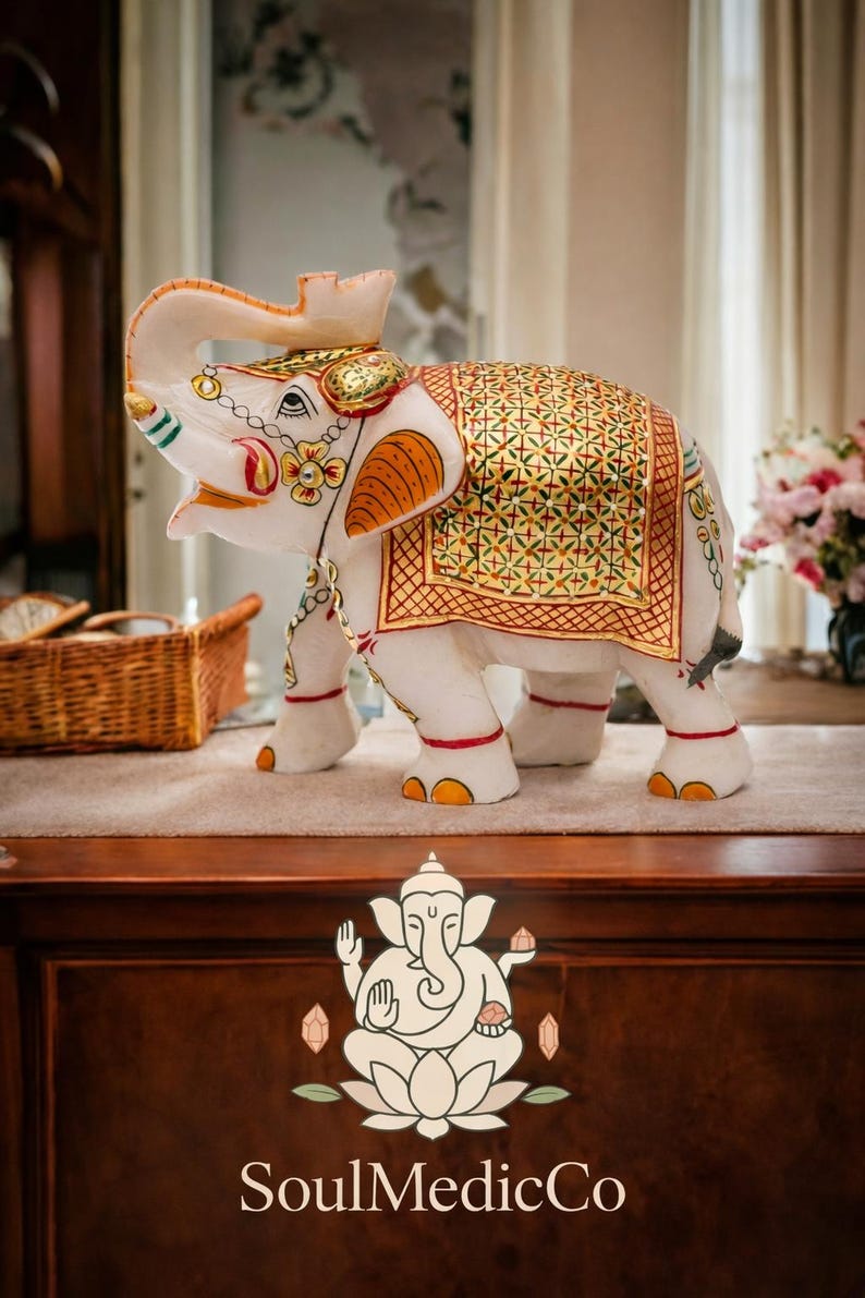 Puede incluir: Una figura de elefante blanco con detalles dorados y naranjas ornamentados. El elefante tiene una manta decorativa con un patr&oacute;n dorado y verde. La figura est&aacute; sobre una superficie de madera. El logo "SoulMedicCo" es visible.