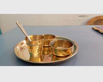 May include: A gold-colored brass serving set, including a round tray, two small bowls, and a cup with a spoon. The set is displayed on a gray surface, reflecting the warm tones of the metal.