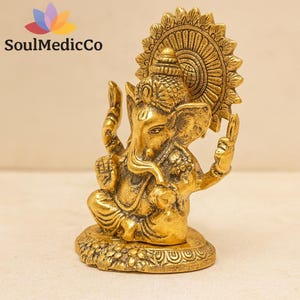 May include: A gold-toned metal figurine of the deity Ganesha, depicted in a seated position with an elephant head, trunk, and four arms. The figure features a large circular halo behind the head and rests on a textured base. Text reads SoulMedicCo.