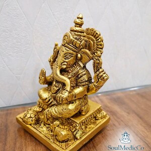 May include: A gold-colored statue of the Hindu deity Ganesha, seated on a square base. The statue features intricate details, including a large head, elephant ears, and multiple arms. The figure is adorned with ornate headdresses and jewelry.