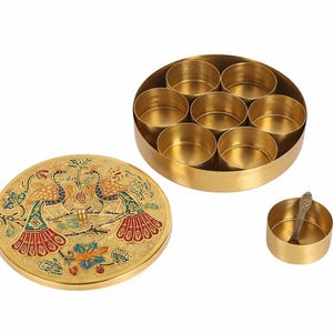 May include: A gold-colored spice box with a lid featuring a peacock design. The box is round with seven compartments, and a small spoon rests in a separate container. The lid is decorated with colorful enamel.