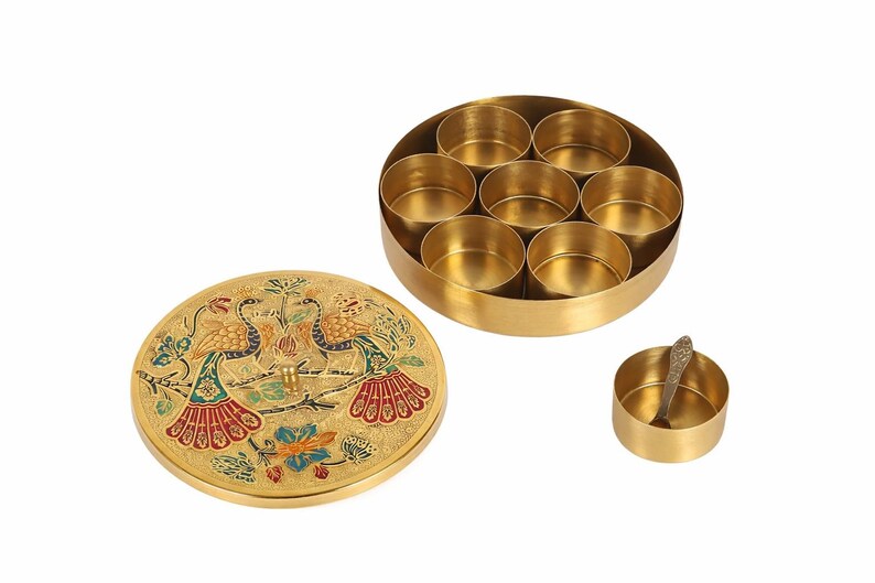 May include: A gold-colored spice box with a lid featuring a peacock design. The box is round with seven compartments, and a small spoon rests in a separate container. The lid is decorated with colorful enamel.
