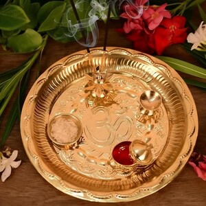 May include: A gold puja thali with detailed patterns, featuring small bowls, a red liquid, and a floral centerpiece. Incense sticks release smoke, and the thali is surrounded by flowers and greenery, creating a spiritual atmosphere.