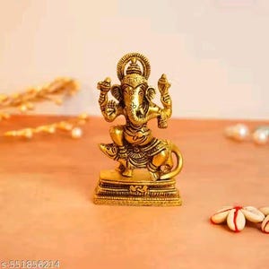 Op de afbeelding: Een goudkleurig beeld van de hindoe&iuml;stische god Ganesha, zittend op een rat. Het beeld heeft ingewikkelde details en wordt tentoongesteld op een vierkante basis. De achtergrond is zacht oranje.