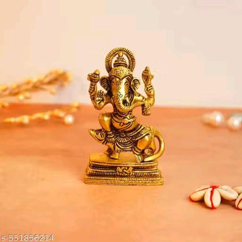 Op de afbeelding: Een goudkleurig beeld van de hindoe&iuml;stische god Ganesha, zittend op een rat. Het beeld heeft ingewikkelde details en wordt tentoongesteld op een vierkante basis. De achtergrond is zacht oranje.