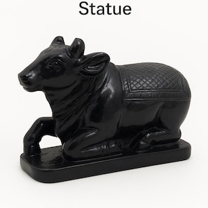 Può includere: Statua in marmo nero lucido di Nandi, il toro, in posizione reclinata. La statua presenta intagli dettagliati sulla sella e si trova su una base rettangolare. Il testo "Marble Shiva Nandi Statue" è in alto.
