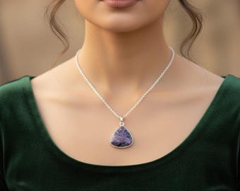 Könnte beinhalten: Eine silberne Halskette mit einem dreieckigen, lila Edelsteinanhänger. Der Anhänger ist in einen silbernen Rahmen gefasst und hängt an einer zarten Silberkette. Die Person trägt ein dunkelgrünes Samt-Oberteil.