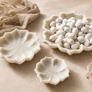 Può includere: Tre ciotole in marmo bianco a forma di fiori di loto con bordi smerlati. La ciotola più grande è riempita con piccoli dolciumi ovali bianchi e lisci. Le ciotole presentano una texture in pietra naturale con venature grigie sottili.