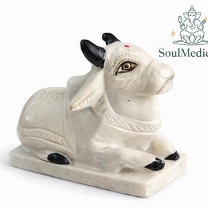Può includere: Statua in marmo bianco di un toro reclinato con corna e zoccoli neri. Il toro presenta lineamenti dettagliati e un punto rosso sulla fronte. La statua è su una base rettangolare, con il logo SoulMedicCo.