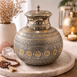 Puede incluir: Un jarrón decorativo plateado con tapa, con intrincados patrones florales y geométricos. El jarrón tiene un cuerpo redondeado y una tapa con un pequeño pomo. Acentos amarillos y blancos contrastan el diseño. El jarrón está sobre una bandeja de mármol.