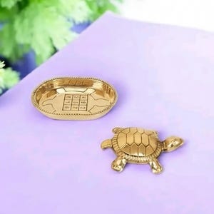 以下が含まれることがあります： 金色の金属製のカメの置物と、格子模様の楕円形のお皿。カメは詳細な甲羅のデザインです。お皿には装飾的な縁と内側に格子模様があります。アイテムは紫色の表面にあります。