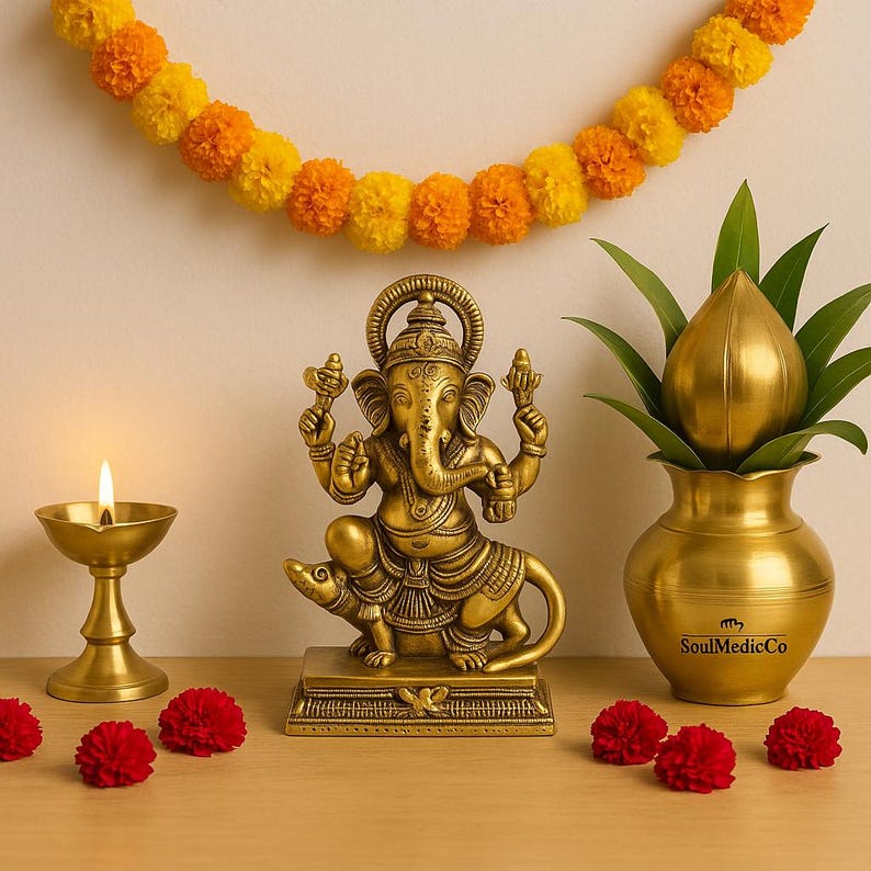 Op de afbeelding: Een gouden messing beeld van de hindoe-god Ganesha, zittend op een rat, met een brandende olielamp en een decoratieve pot met een lotusknop. Op de achtergrond een slinger van oranje en gele bloemen en rode anjers.