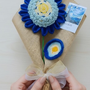 Może przedstawiać: Ręcznie robiony bukiet kwiatów na szydełku z dużym niebieskim i jasnoniebieskim kwiatem z żółtym środkiem. Bukiet jest owinięty w jutę i zawiązany wstążką. Dołączona jest mała karta z obrazem.