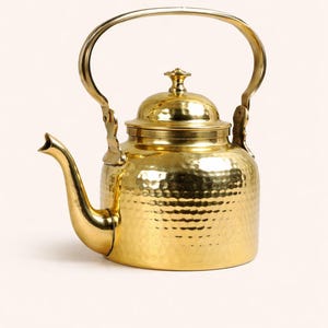 May include: A hammered brass teapot with a curved spout and a large handle. The teapot has a lid with a small decorative knob. The overall design is a classic, vintage style. The teapot measures approximately 8 inches in height.
