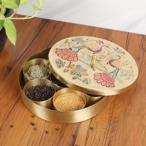 May include: A gold-colored spice box with a decorative lid featuring a peacock design. The box is open, revealing several compartments filled with various spices. The box sits on a wooden surface.