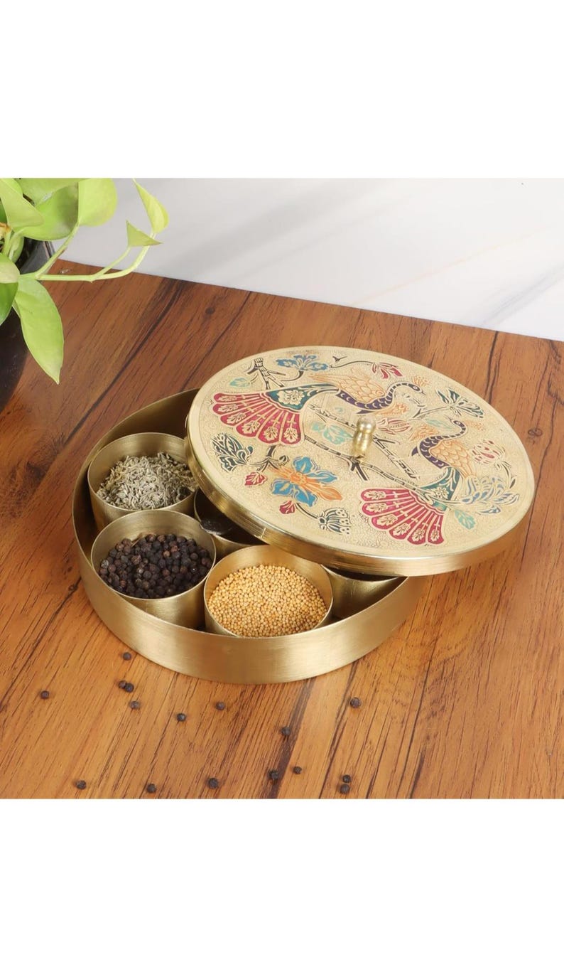 May include: A gold-colored spice box with a decorative lid featuring a peacock design. The box is open, revealing several compartments filled with various spices. The box sits on a wooden surface.