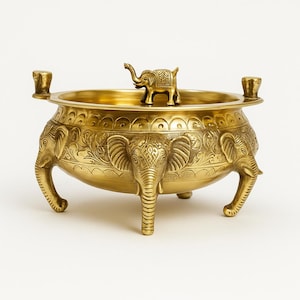 May include: A gold-colored, decorative bowl with an elephant motif. The bowl has three elephant-shaped legs and three elephant heads around the sides. A small elephant figurine sits in the center. The bowl has two small handles.