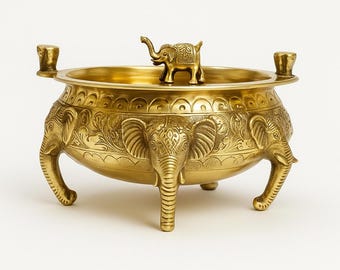 Puede incluir: Un cuenco decorativo de color dorado con un motivo de elefante. El cuenco tiene tres patas con forma de elefante y tres cabezas de elefante en los lados. Una pequeña figura de elefante se encuentra en el centro. El cuenco tiene dos asas pequeñas.