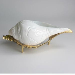Könnte beinhalten: Eine weiße, muschelförmige Dekoschale mit einem goldfarbenen Metallsockel. Die Schale hat eine strukturierte Oberfläche und wird von drei kleinen Beinen getragen. Der goldene Sockel hat ein blattartiges Design.