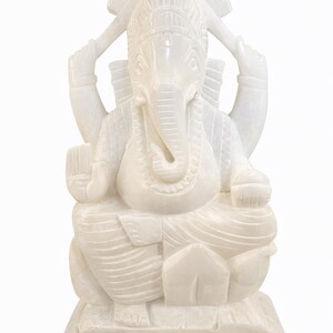 Può includere: Statua in marmo bianco di Ganesha, divinità induista, seduta a gambe incrociate. La scultura presenta dettagli intricati, tra cui una corona, una testa di elefante e più braccia che tengono vari oggetti. La statua è su una base quadrata.